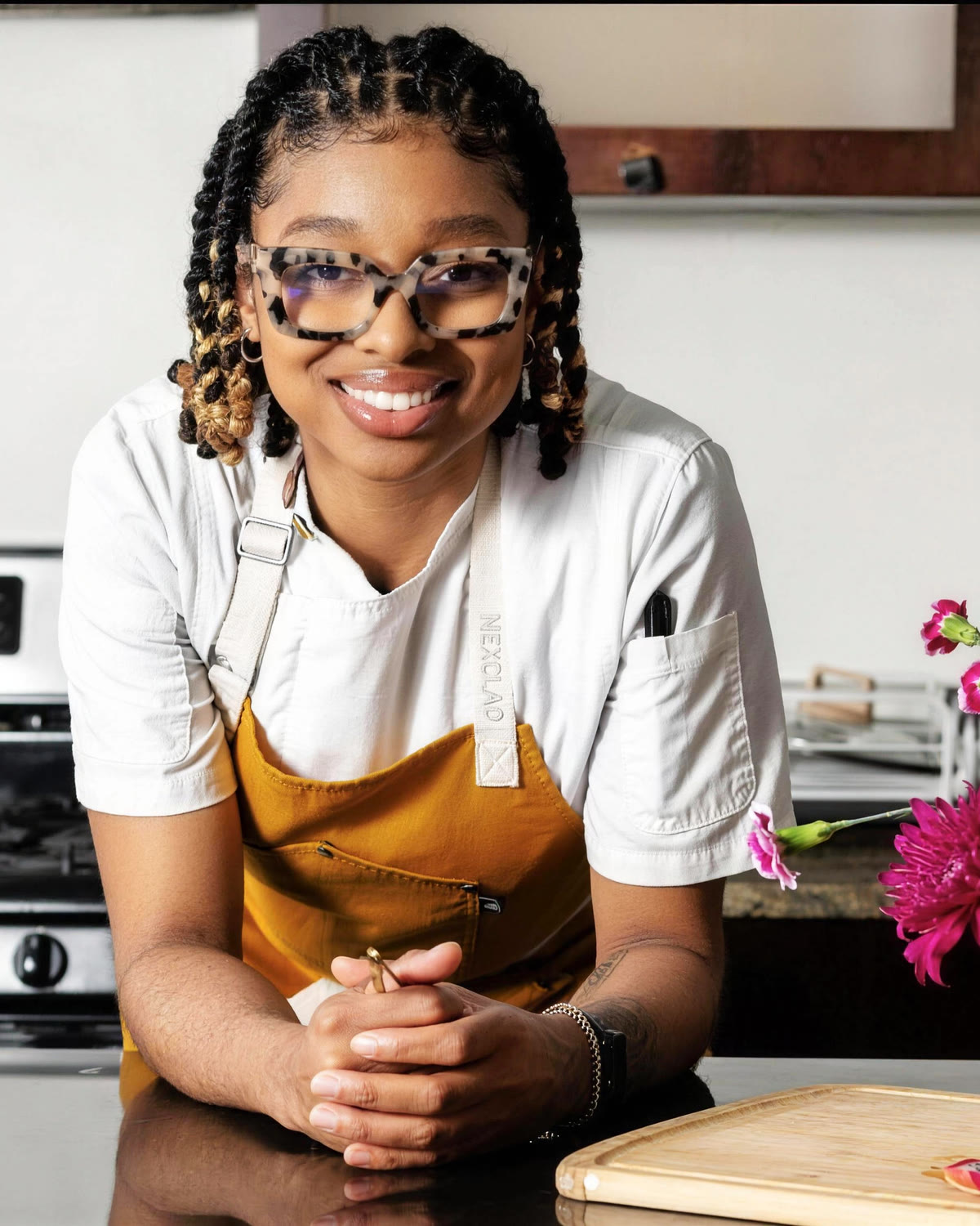 Chef Nia Spencer leaning on a kitchen island in her whites and apron, beside a bright flower arrangement