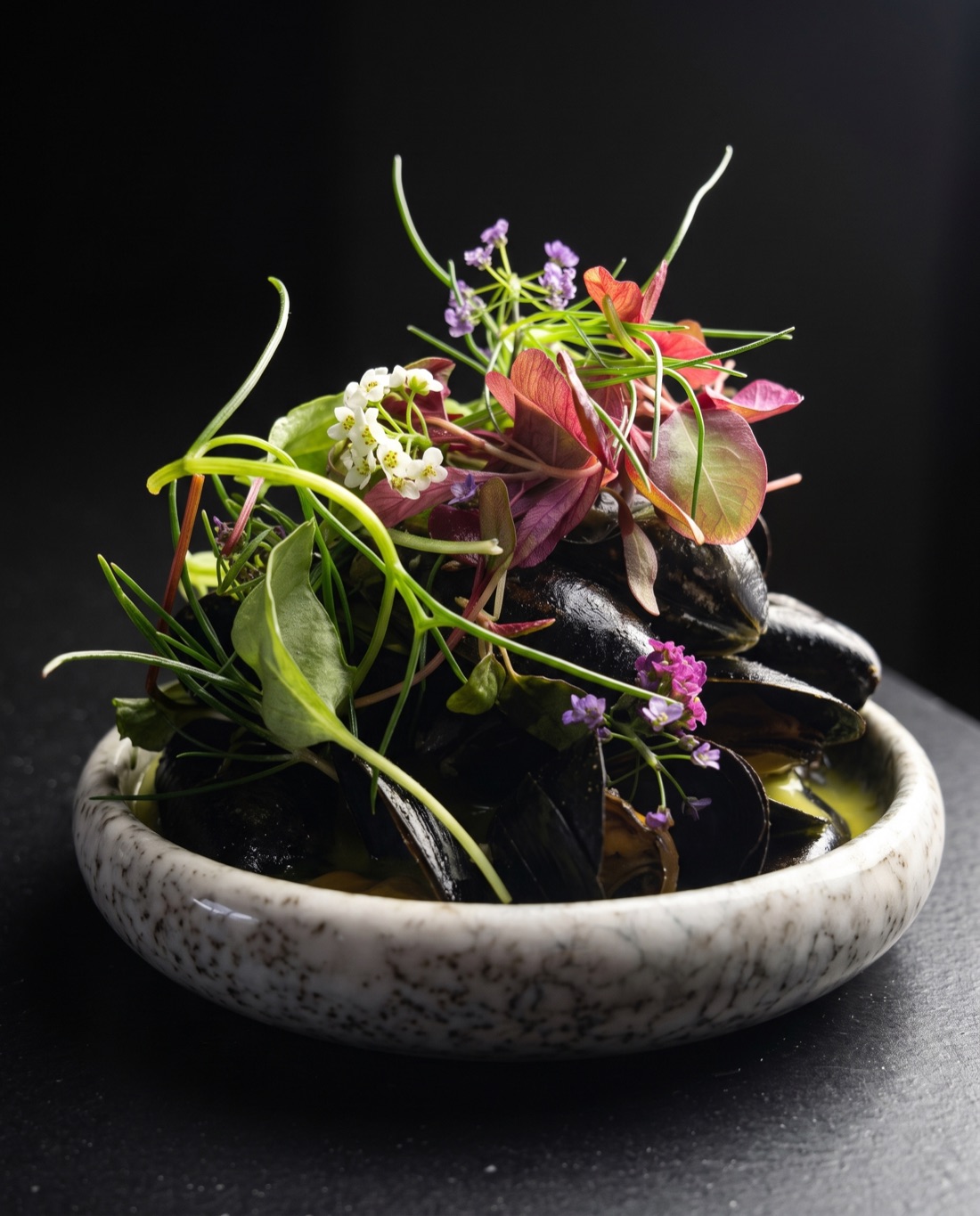 Mussels in green herb broth, the crown of flowers and micro herbs rim-lit against a dark background