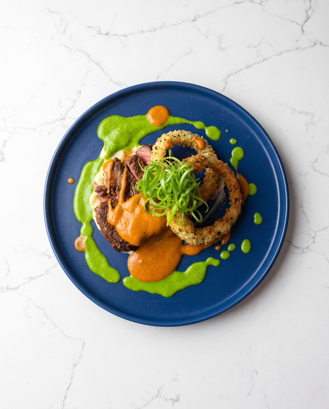 Braised short rib with romesco, herb purée and a panko-crusted onion ring on cobalt, seen from directly above